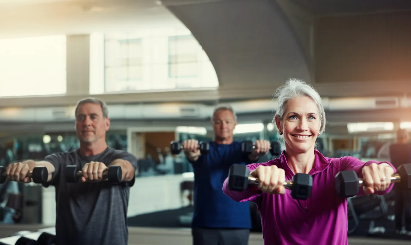 Trois adultes plus âgés, une femme aux cheveux blancs souriante vêtue d’un haut magenta et deux hommes, effectuant ensemble des élévations d’haltères debout dans un gymnase lumineux.