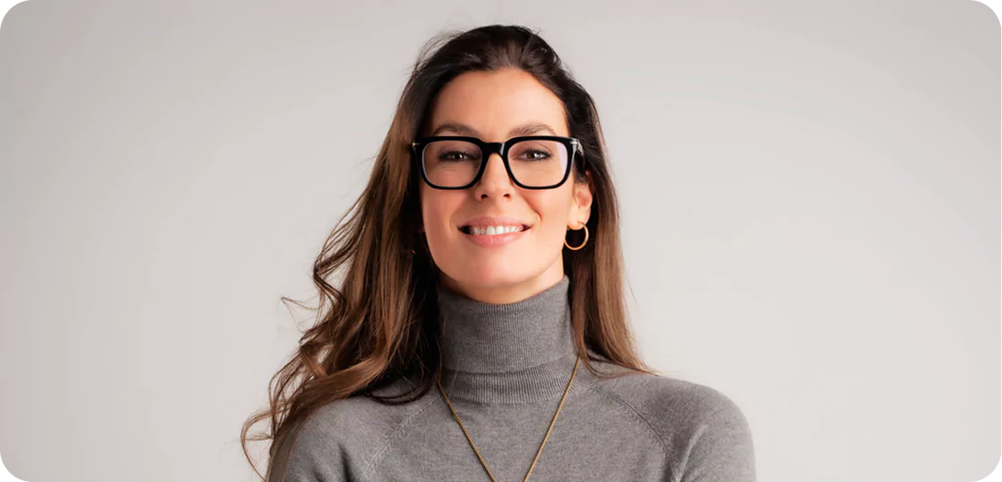 Confident brunette woman with crossed arms, wearing glasses and a sweater, standing against a plain background while looking directly at the camera.