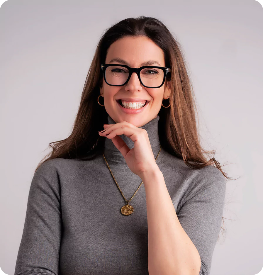 Close-up of a confident mid-aged woman in a turtleneck and glasses, set against a plain grey background.