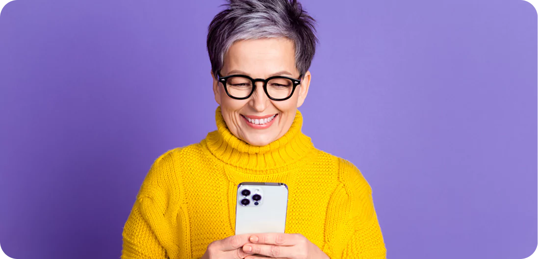 Middle-aged woman in yellow knitwear browsing on her phone, looking down, against a violet background.