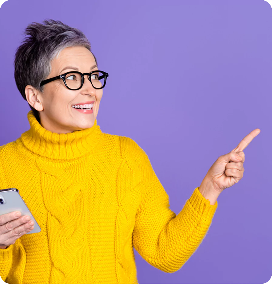Middle-aged woman in yellow knitwear holding a phone and pointing right, posed against a violet background.