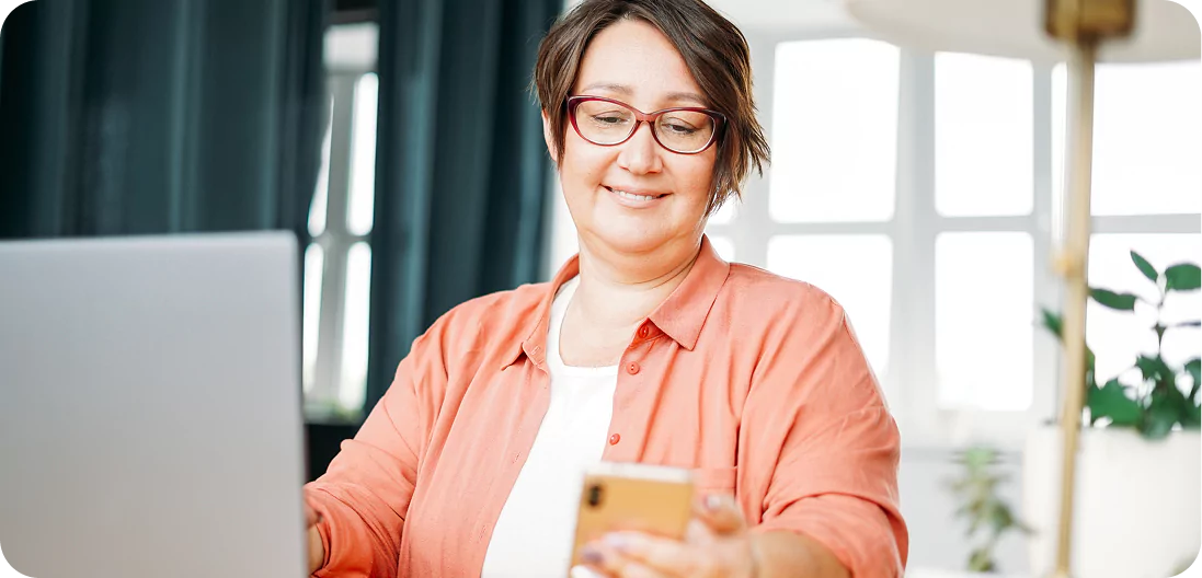 Smiling plus-size brunette woman in glasses, seated with a laptop in front of her, glancing at a smartphone off to the side.
