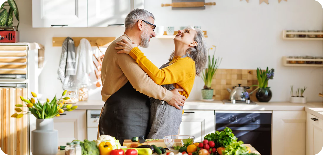 Joyful senior couple smiling and embracing while cooking together in the kitchen, preparing vegetarian dishes.