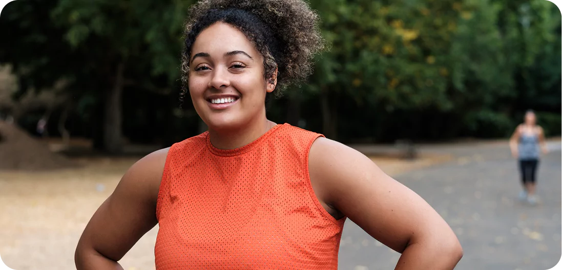 Smiling young woman with curly hair standing on a park road post-jog, wearing a sleeveless orange t-shirt — proud, confident, and triumphant.