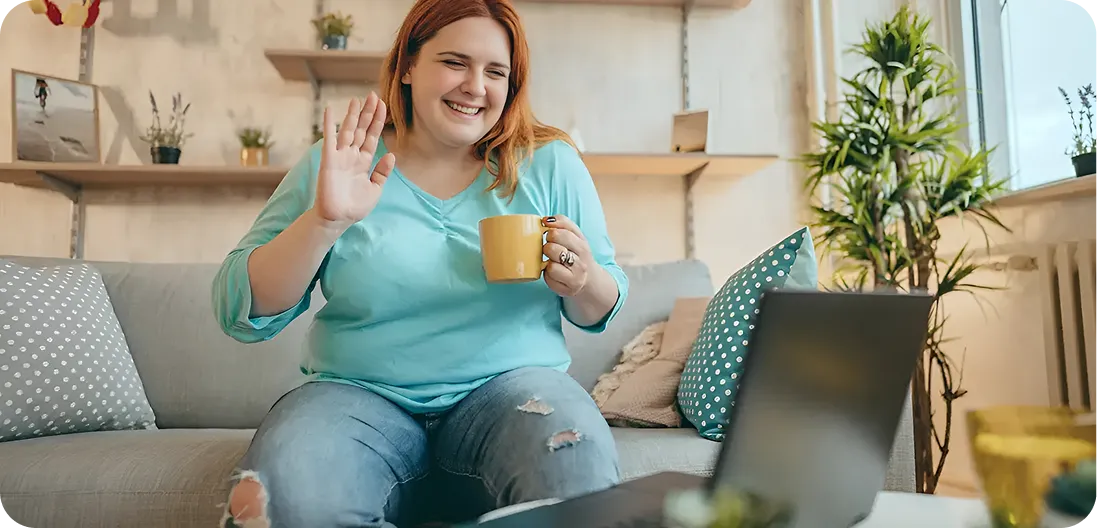 Smiling young red-haired woman sitting on a sofa, waving at her laptop with one hand and holding a yellow coffee cup in the other — relaxed, mid-video call with her dietitian.